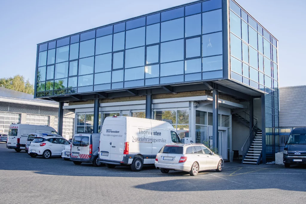 Gebäude mit verglastem Obergeschoss, davor Transporter und Pkw, darunter ein Transporter mit der Aufschrift Thorwesten Nutzfahrzeugservice